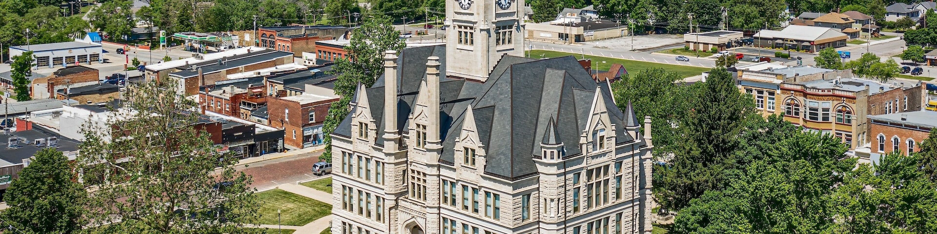 An aerial view of a historic building with a clock tower, surrounded by greenery and urban structures.