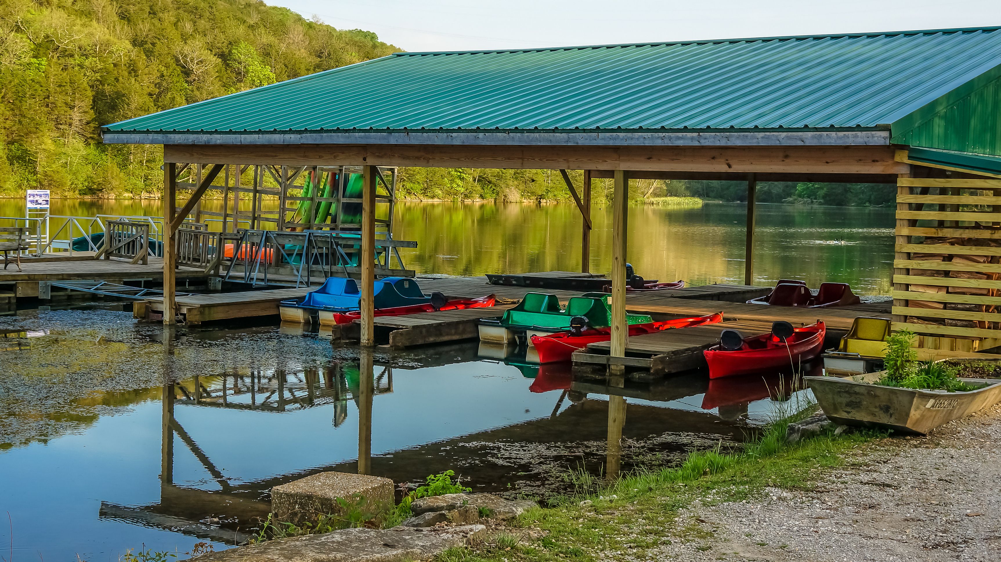 A lakeside dock with boats, a covered pavilion, and a forested hill in the background.