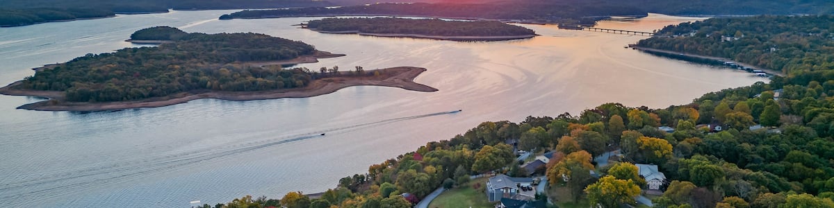 Un coucher de soleil sur une rivière parsemée d’îles, entourée d’une forêt dense et de zones résidentielles.