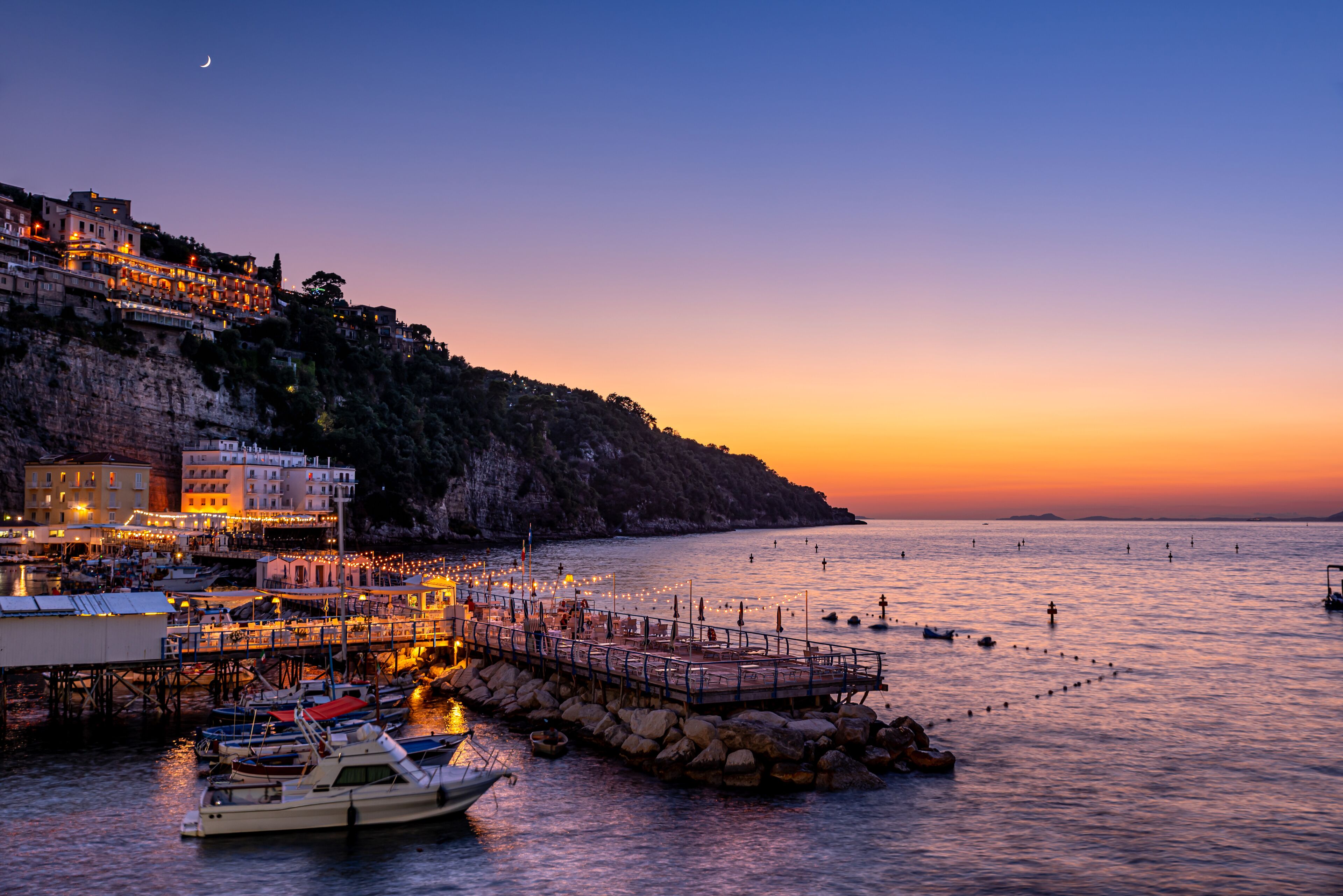 Uma vila costeira ao pôr do sol, com edifícios em um penhasco, um cais com barcos e mar calmo.