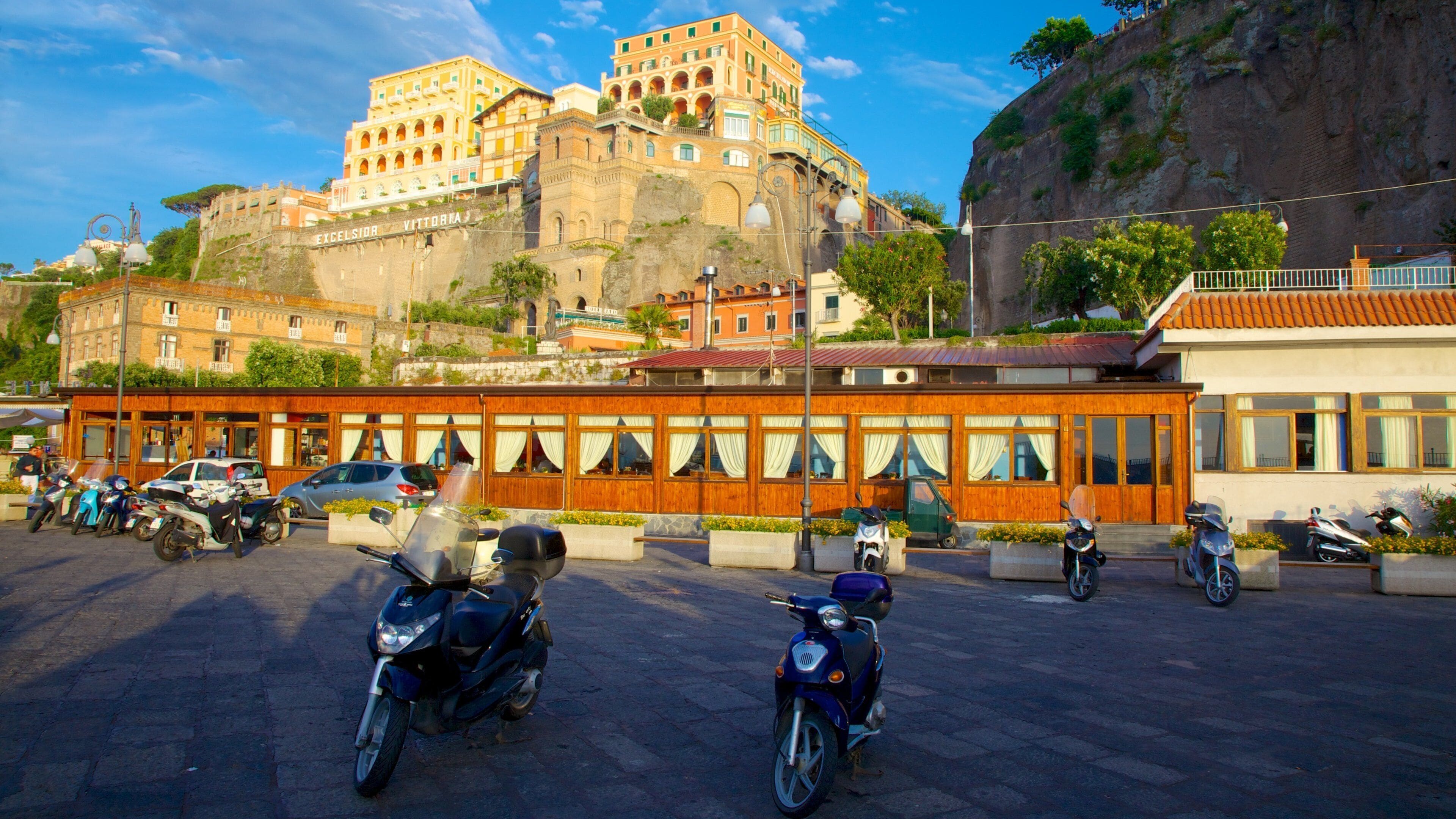 A historic building with a stone facade and multiple balconies, surrounded by modern buildings and parked scooters.