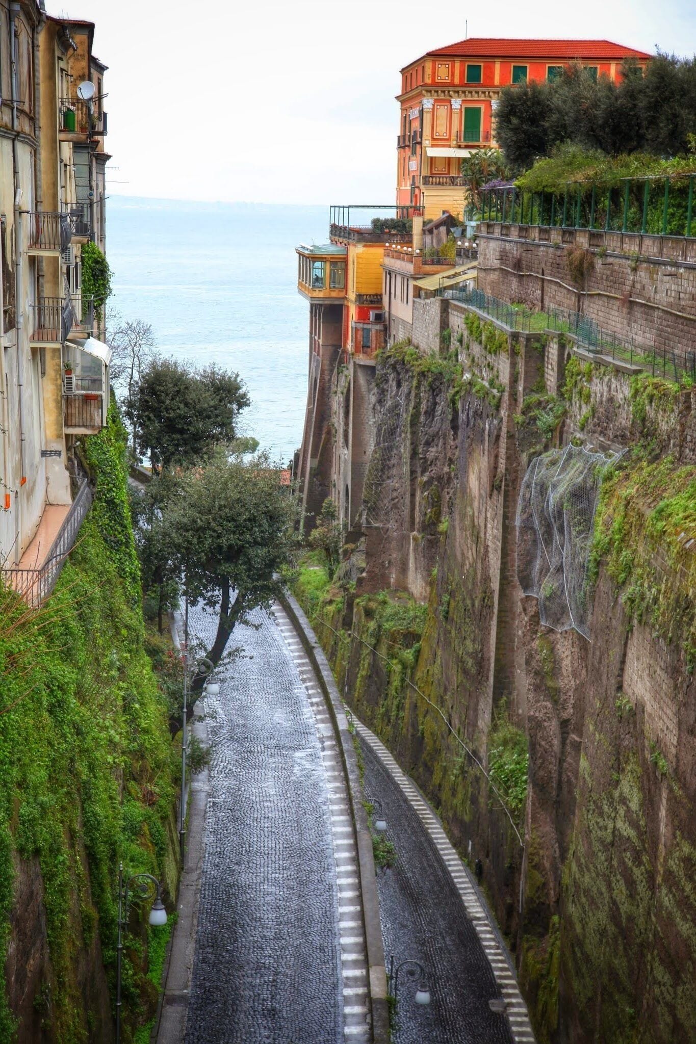 A narrow cobblestone street flanked by steep cliffs and buildings with balconies overlooking a body of water.