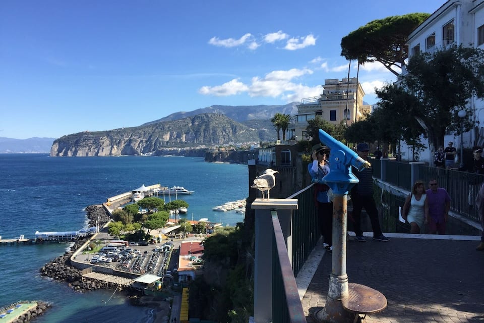 A coastal town with a clear view of the sea, a seagull perched on a railing, and a building with a balcony overlooking the water.