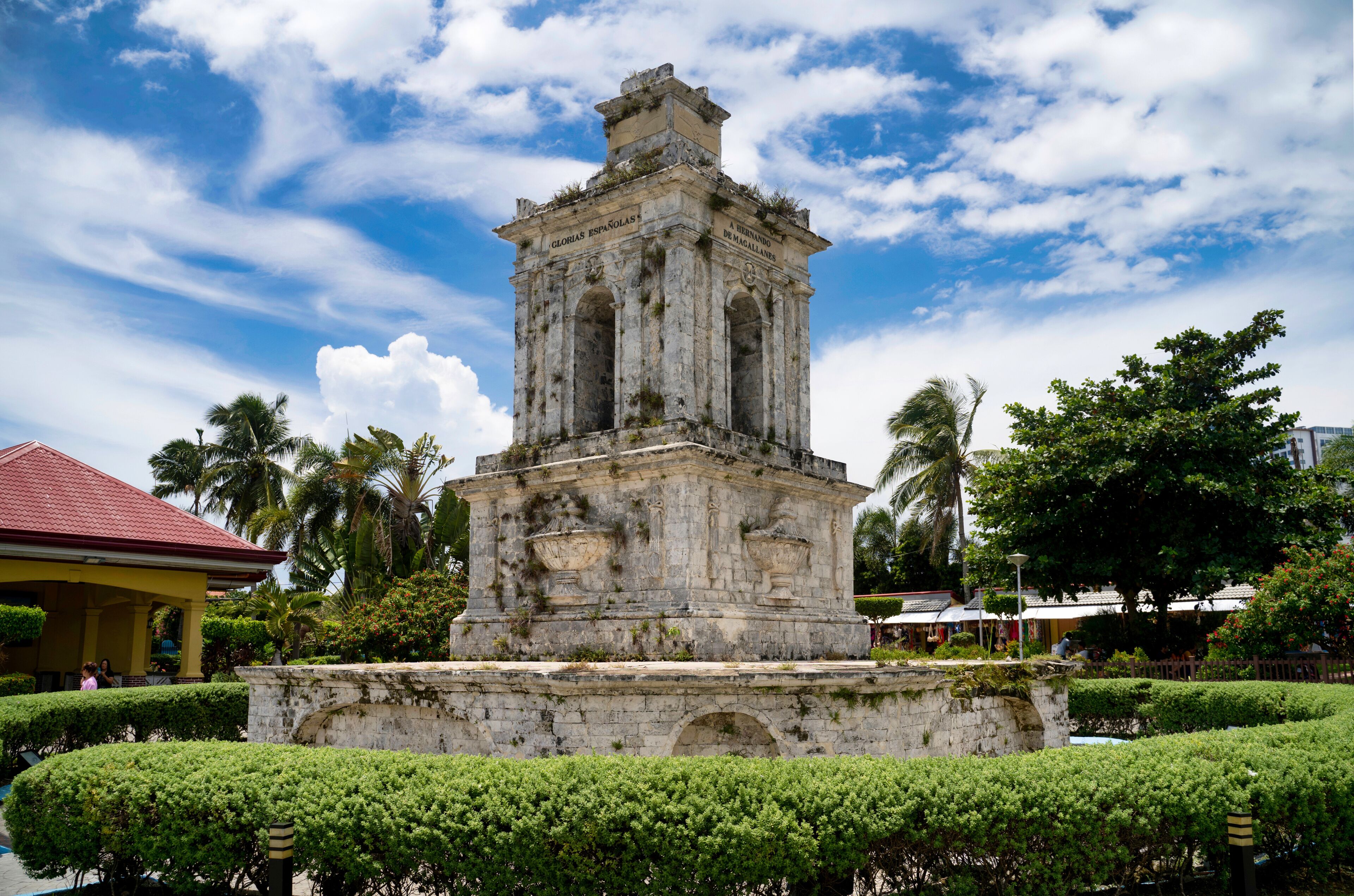 A historical monument with inscriptions, surrounded by well-maintained hedges and palm trees.