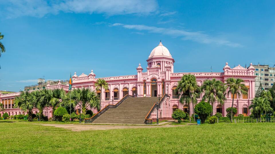 A pink colonial-style building with a dome and steps leading up to it, surrounded by palm trees and a grassy field.