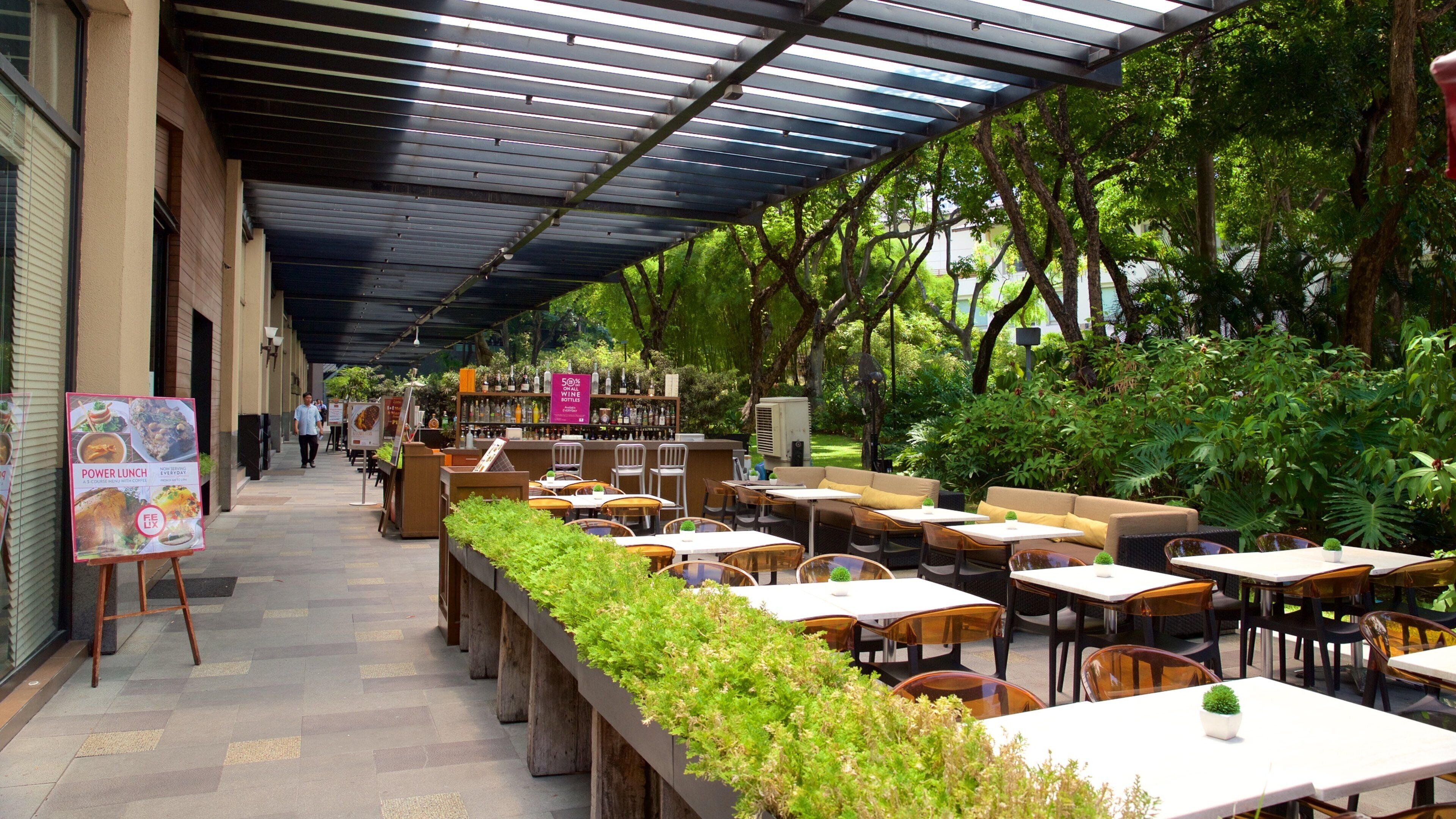 An outdoor café with tables and chairs, surrounded by greenery and a covered walkway.