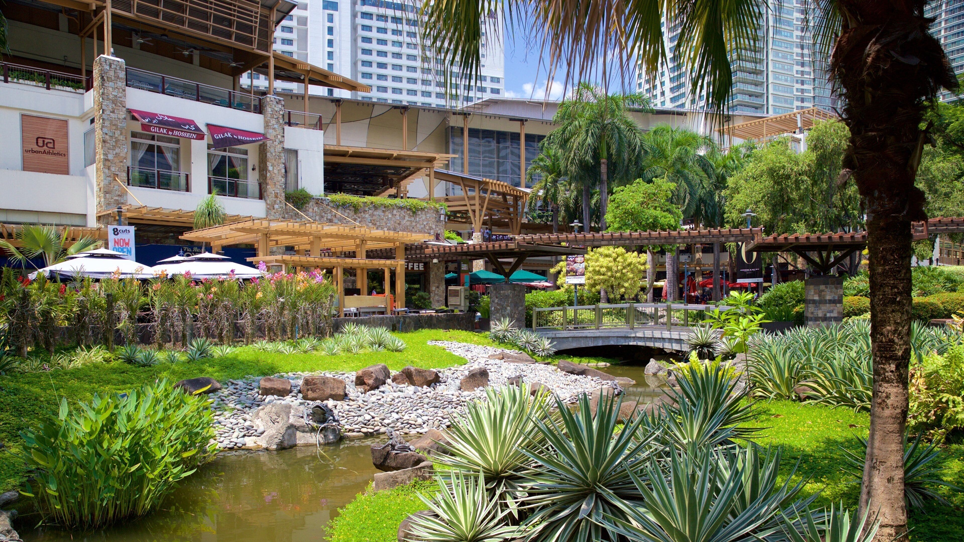 A lush garden with a small pond, a wooden bridge, and a restaurant named Gilak & Hossein.