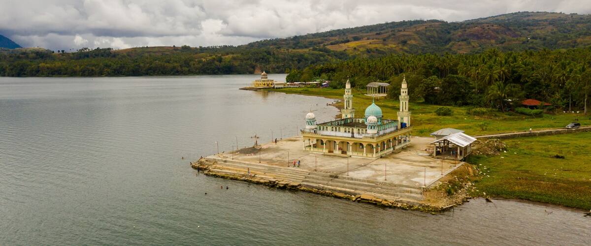 A mosque with a large dome and minarets on an island in a lake, surrounded by hills and palm trees.