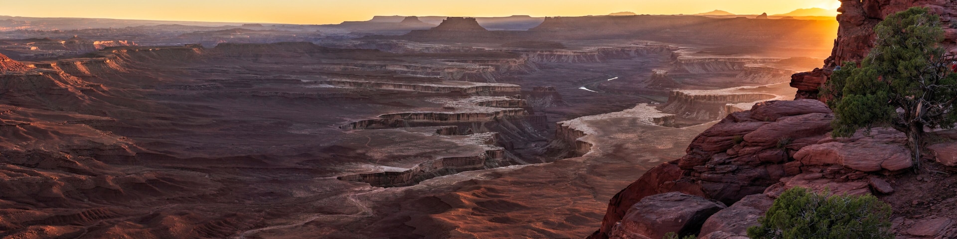 Un extenso paisaje cañonero con formaciones rocosas estratificadas y un atardecer al fondo.