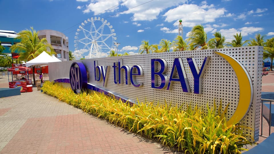 A sign with 'By the BAY' written on it, a Ferris wheel, palm trees, and a building in the background.