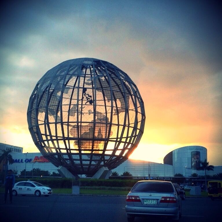 A large spherical structure with a grid pattern, set against a sunset sky, with a building labeled "Hall of Asia" in the background.