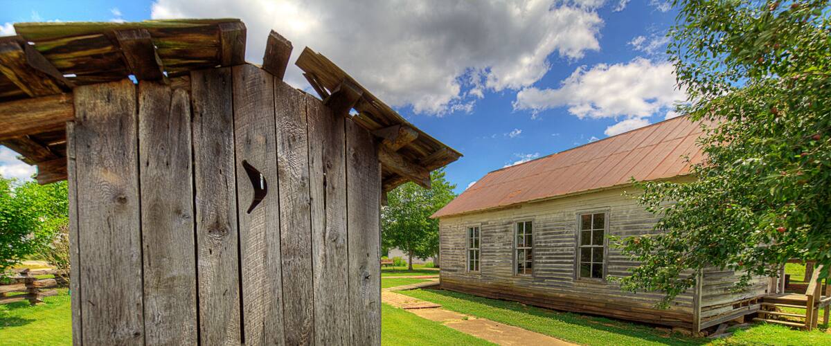 A rustic wooden structure with a metal roof, a wooden fence, and a building with a red roof in a grassy area.