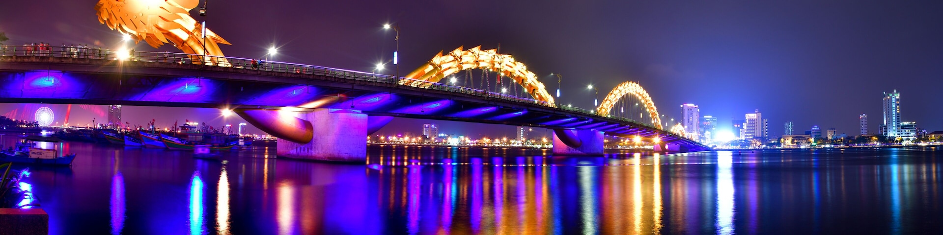 Eine Brücke mit beleuchteten Bögen über dem Wasser, das die Lichter der Stadt spiegelt.