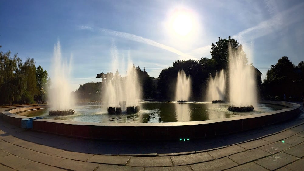 A circular fountain with multiple jets of water, surrounded by a paved walkway and trees.