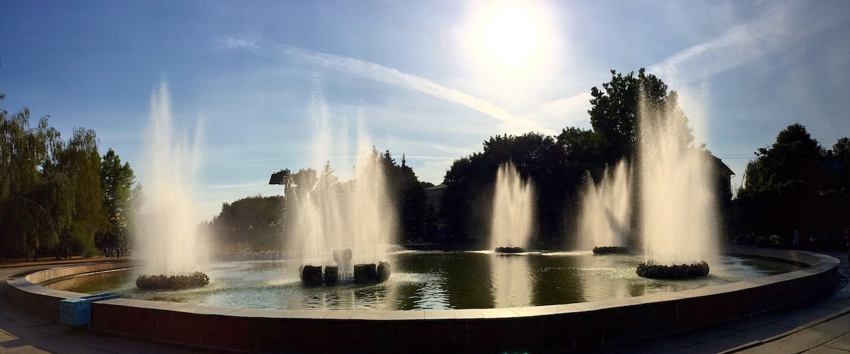 A circular fountain with multiple jets of water, surrounded by a paved walkway and trees.