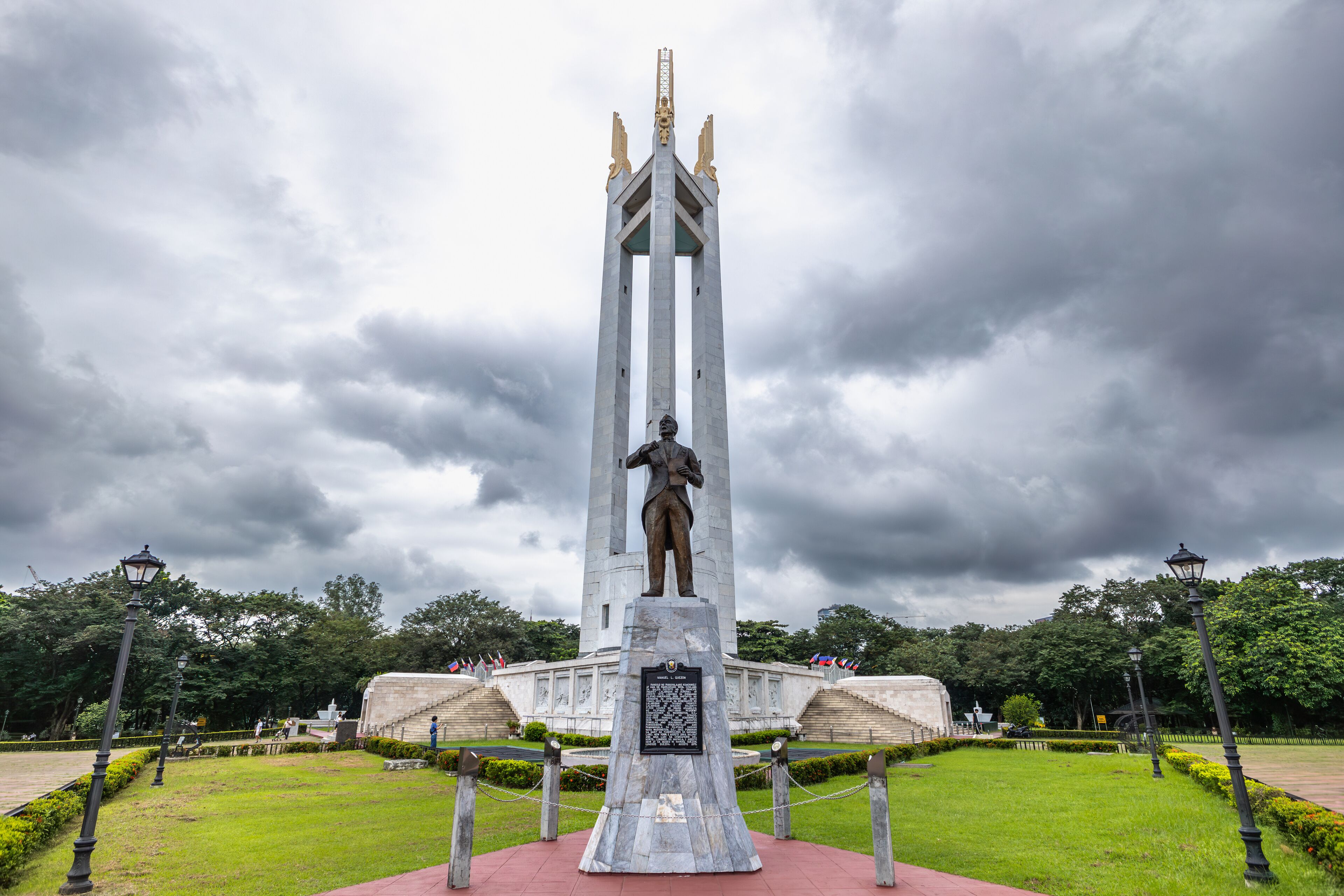 A monument with a statue, surrounded by a park with grass and trees.