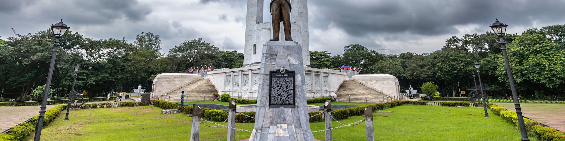 A monument with a statue, surrounded by a park with grass and trees.