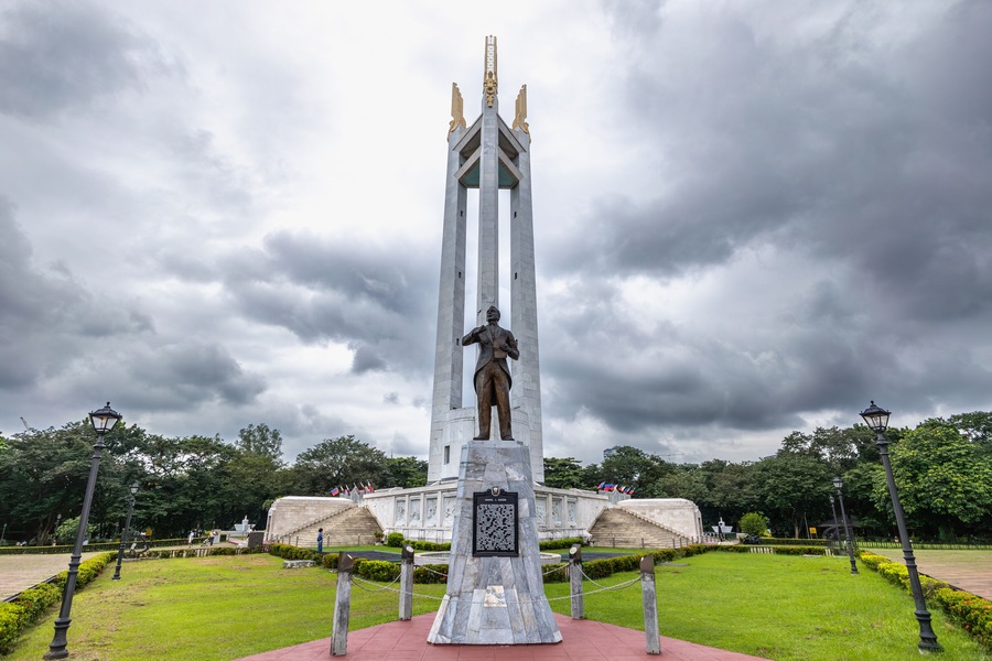 A monument with a statue, surrounded by a park with grass and trees.