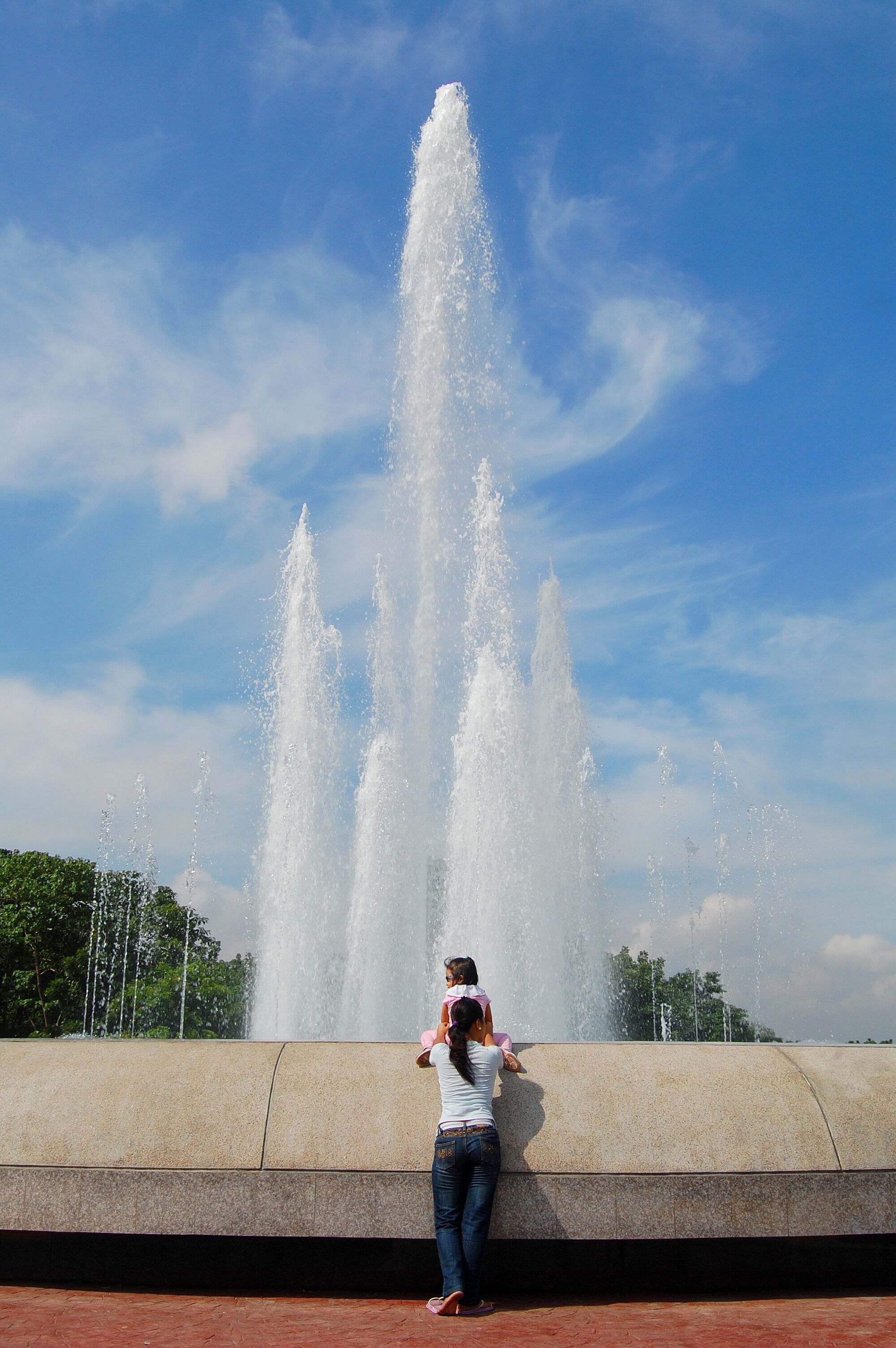 A woman taking a photo of a fountain with multiple water jets against a clear blue sky.