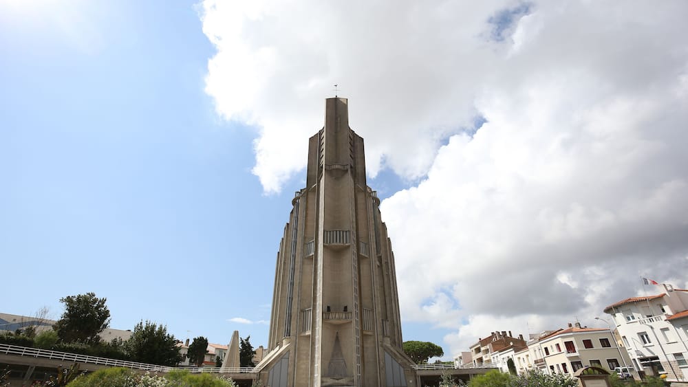 Un grand bâtiment de style moderniste, doté d’une flèche centrale, et entouré d’un jardin bien entretenu où poussent diverses plantes et fleurs.