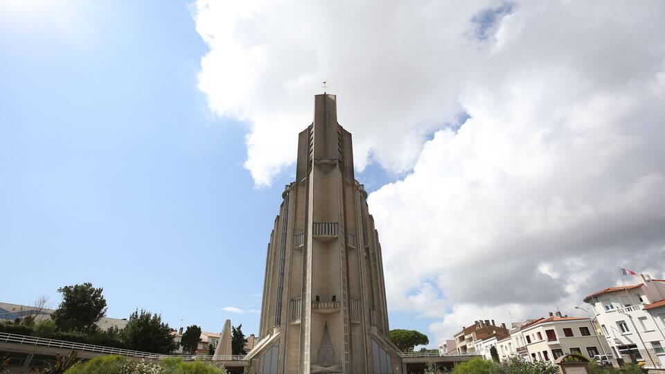 A tall, modernist building with a central spire, surrounded by a well-maintained garden with various plants and flowers.