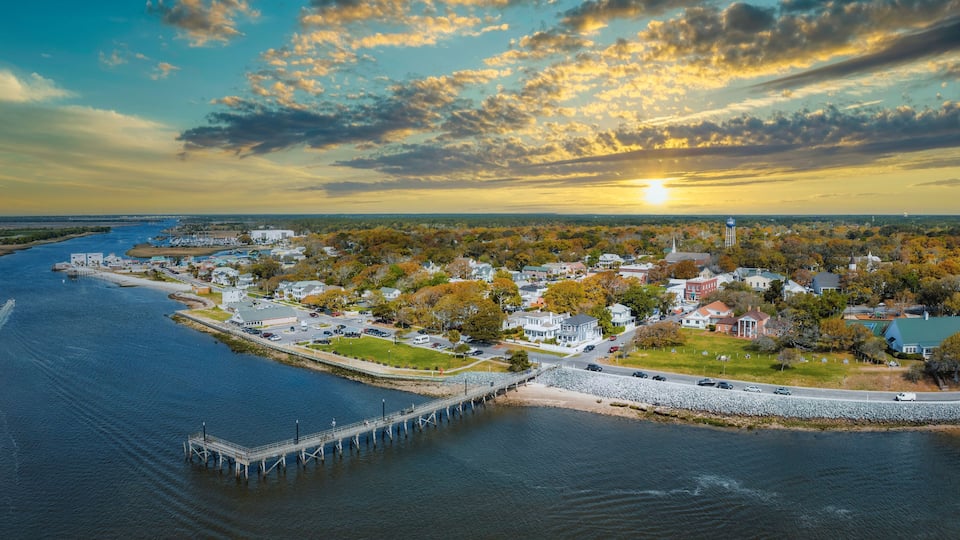 A coastal town with a pier extending into the water, surrounded by autumn-colored trees and a sunset in the background.