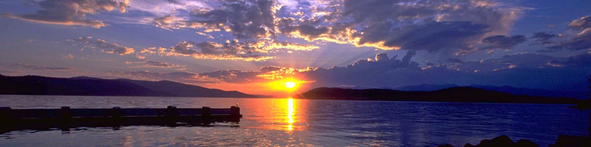 A sunset over a calm lake with a wooden dock and mountains in the background.