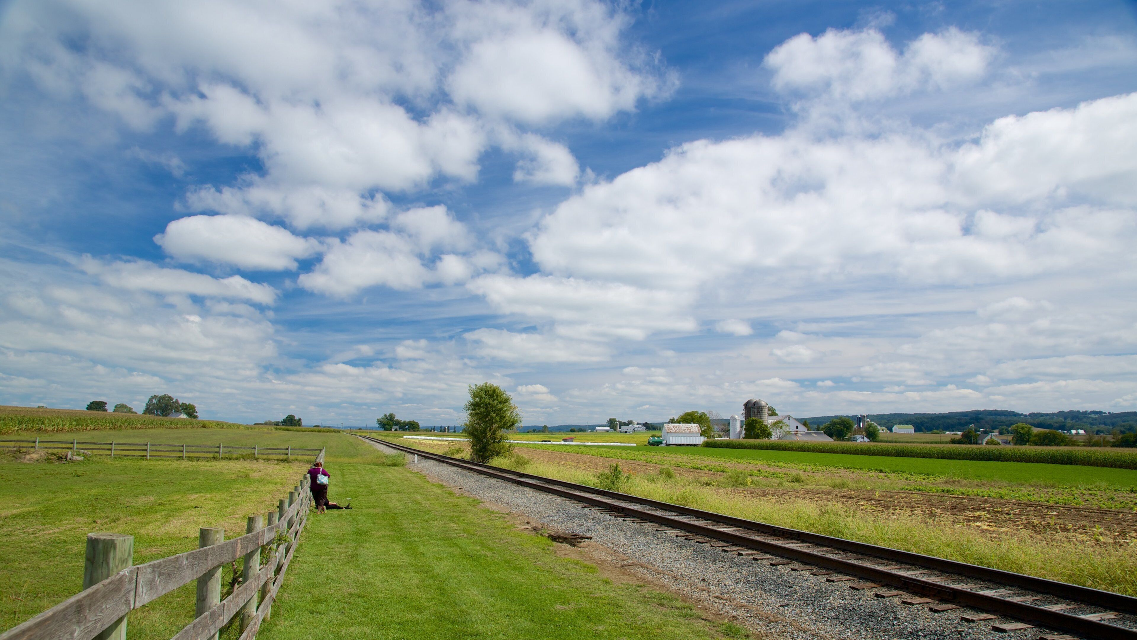 A rural landscape with a railway track, green fields, a wooden fence, and a partly cloudy sky.