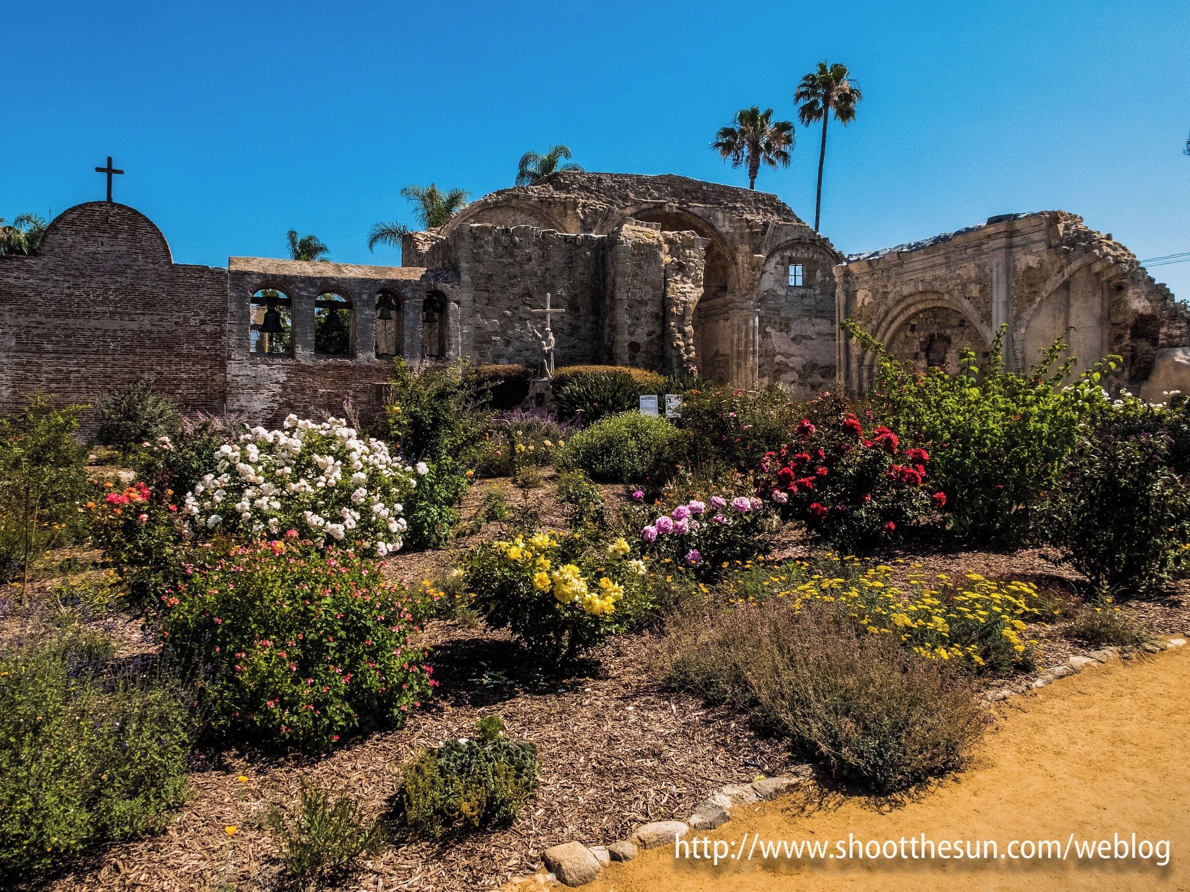 An ancient stone structure with a cross, surrounded by colorful gardens and palm trees.