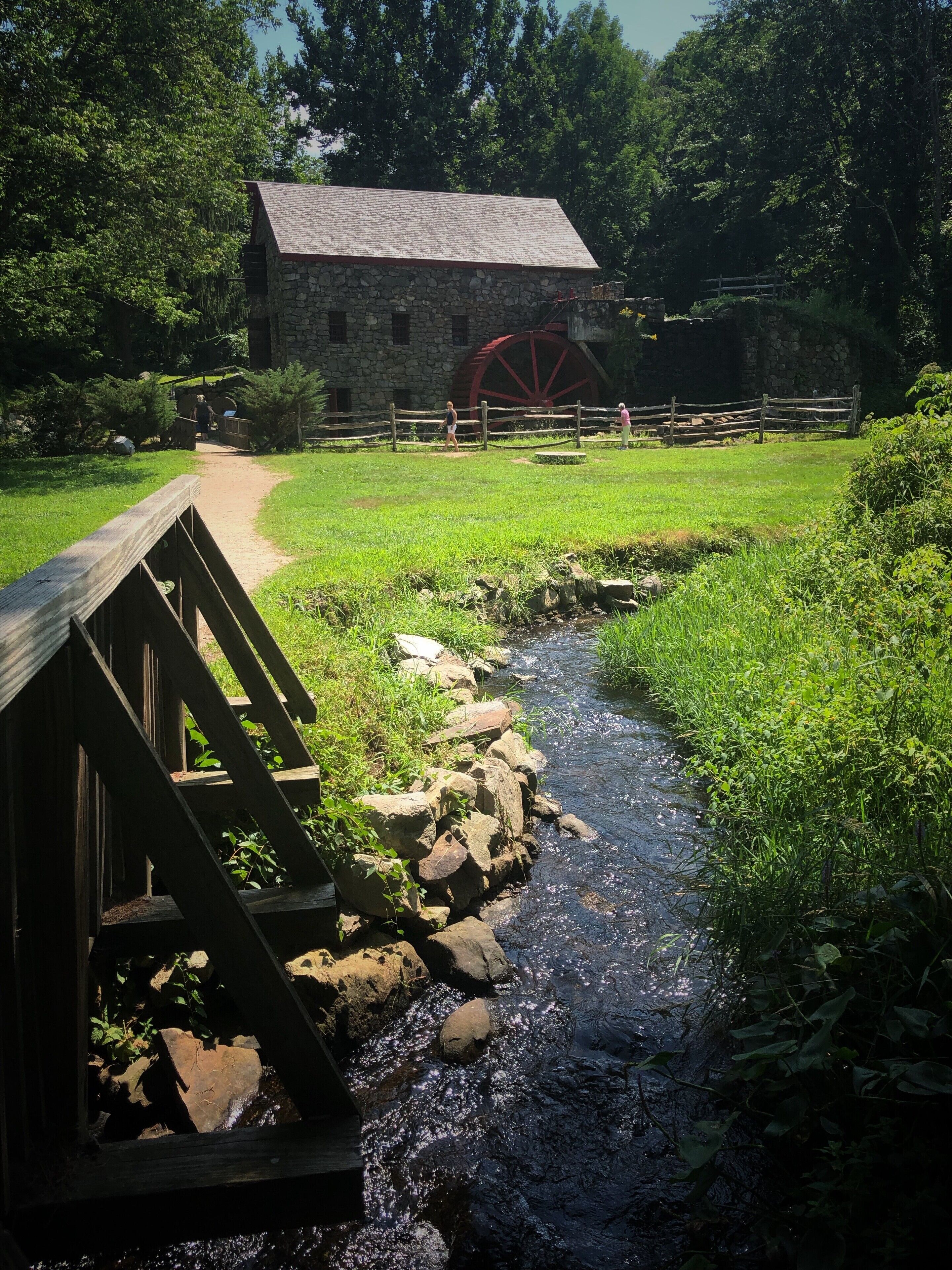 A wooden bridge over a stream, a stone building, and a large red water wheel.