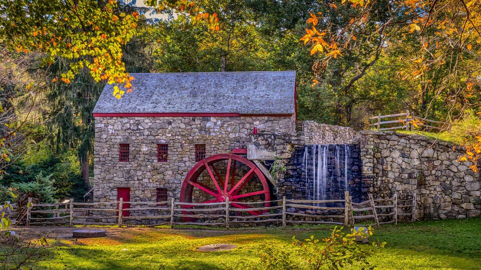 A stone mill with a large red water wheel, surrounded by lush greenery and a wooden fence.