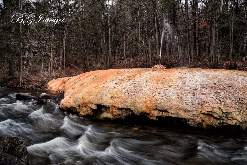 A river flowing over a large, orange-brown rock formation surrounded by a forest.