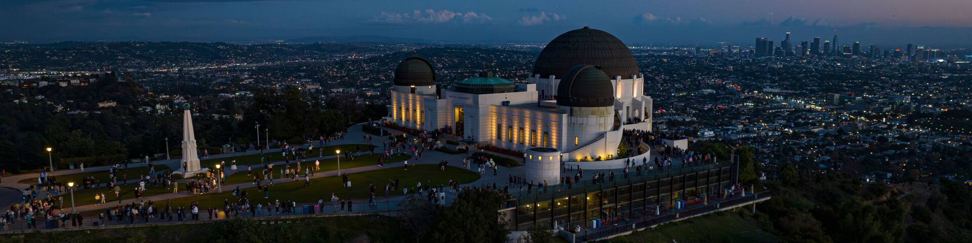 A large observatory with two domes, surrounded by a park with people, set against a cityscape at dusk.