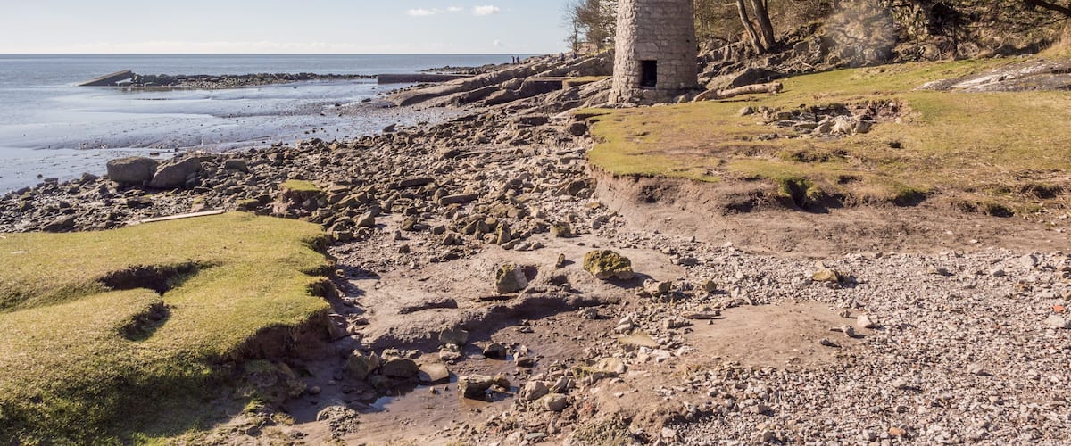 Paesaggio costiero con una torre di pietra, spiaggia rocciosa e cielo sereno.