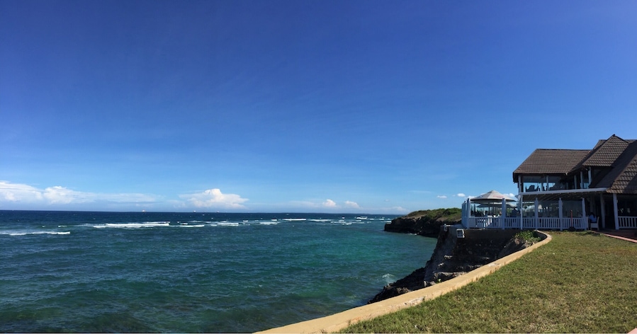 A coastal view with ocean waves, a grassy area, and a house with a balcony overlooking the sea.
