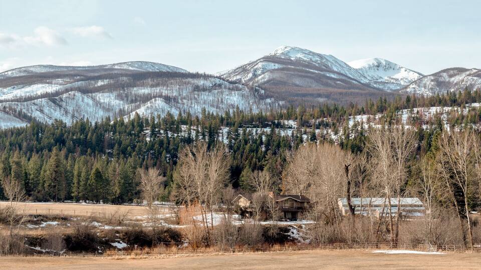 A mountainous landscape with snow-covered peaks, dense forests, and a few buildings in the foreground.