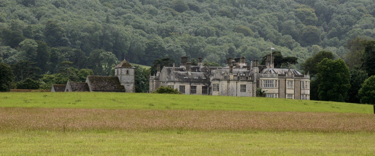 A large, historic stone building surrounded by lush green fields and a dense forested hill in the background.