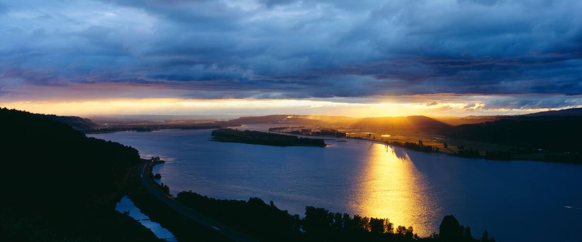 A sunset over a lake with surrounding hills and a road curving along the water's edge.