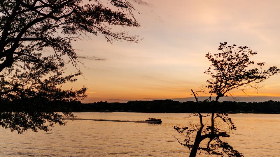 A sunset over a calm lake with a boat in the distance and trees framing the scene.