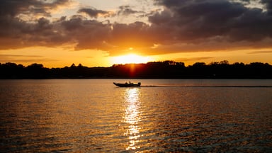 Een zonsondergang boven een rustig meer, met een boot op de voorgrond en een silhouet van bomen aan de horizon.