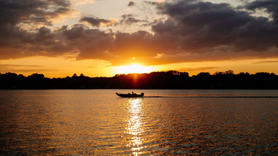 A sunset over a calm lake with a boat in the foreground and a silhouette of trees on the horizon.