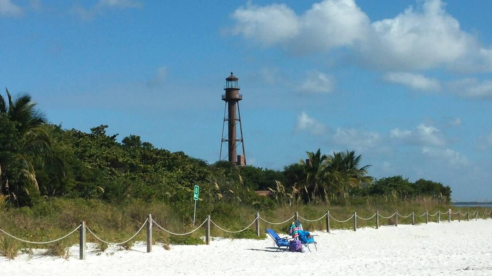 A beach with a lighthouse, palm trees, and a chain-link fence.