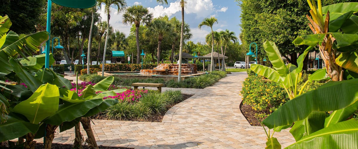 A paved pathway lined with tropical plants and palm trees, leading to a gazebo and a fountain.