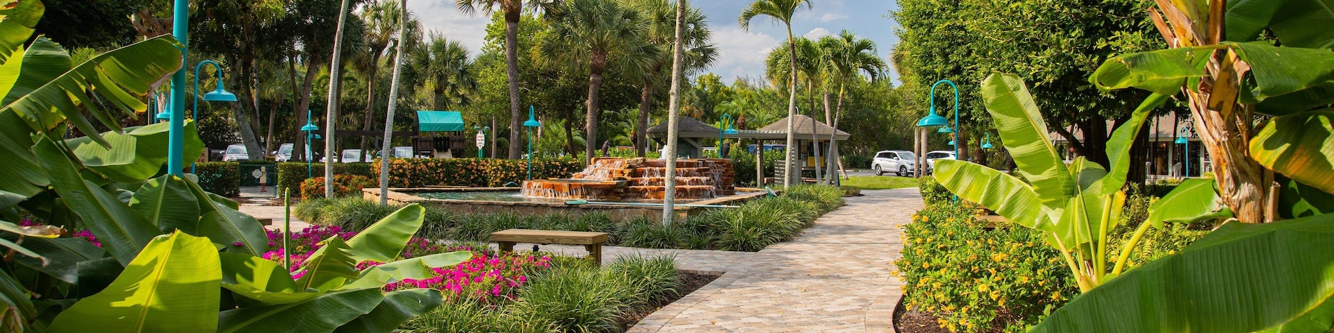 A paved pathway lined with tropical plants and palm trees, leading to a gazebo and a fountain.