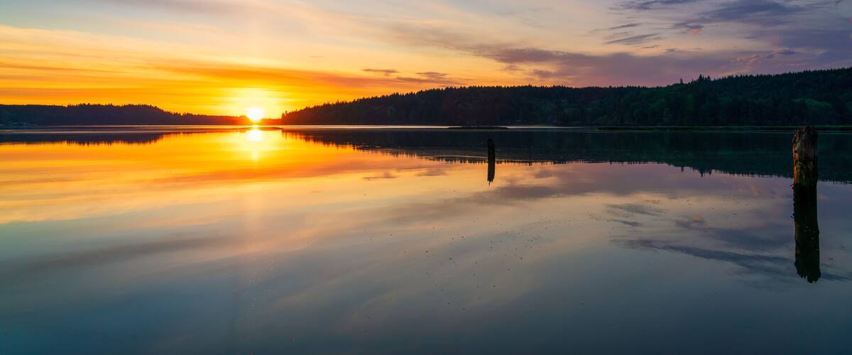 A calm lake at sunset with a forested shoreline and a clear reflection of the sky.
