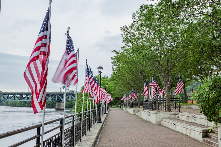 A river with a bridge, a walkway lined with American flags, and trees.