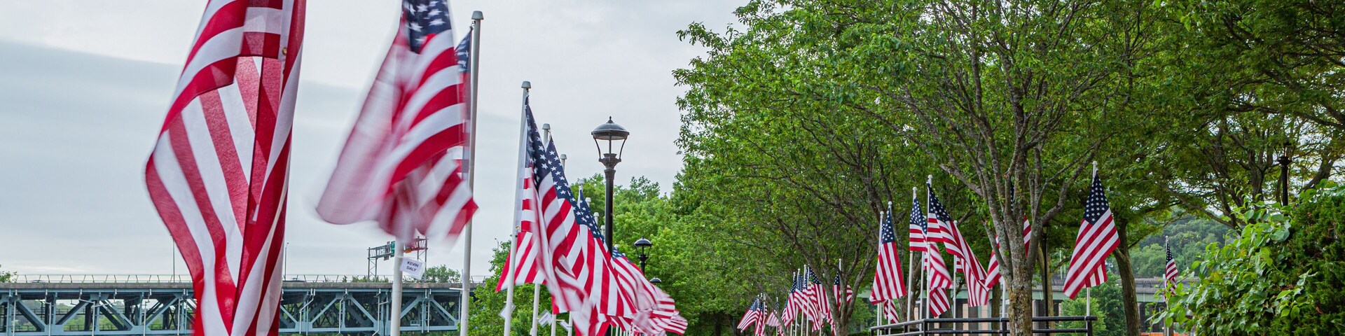 A river with a bridge, a walkway lined with American flags, and trees.