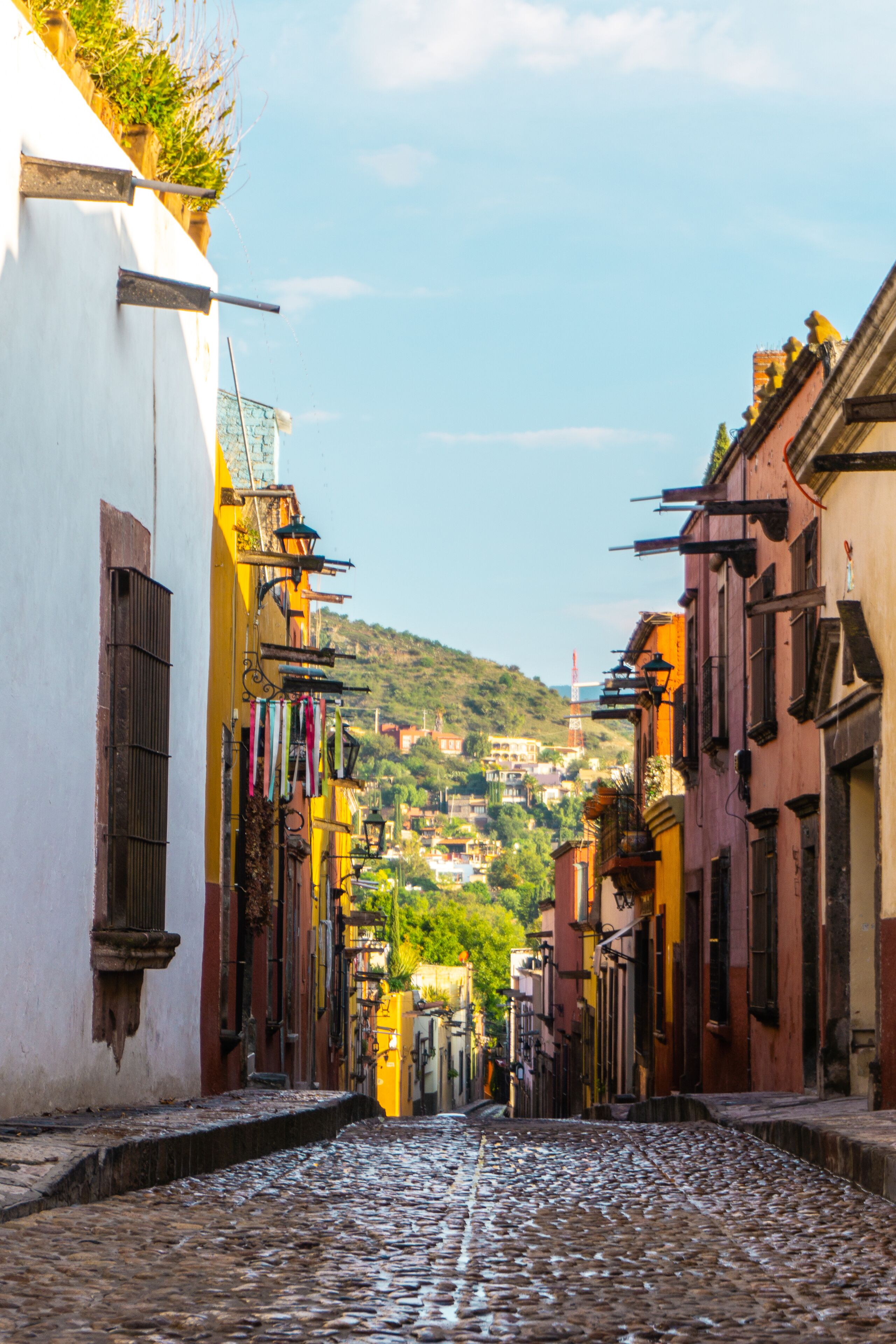 Una calle empedrada bordeada por edificios de colores, una colina con vegetación y una torre distante.