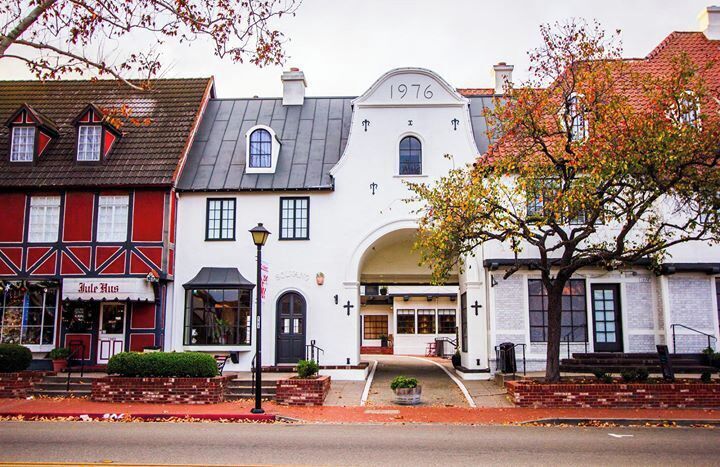 A white building with '1976' on the facade, a red building with 'Jule Hus' signage, and a tree with autumn leaves.