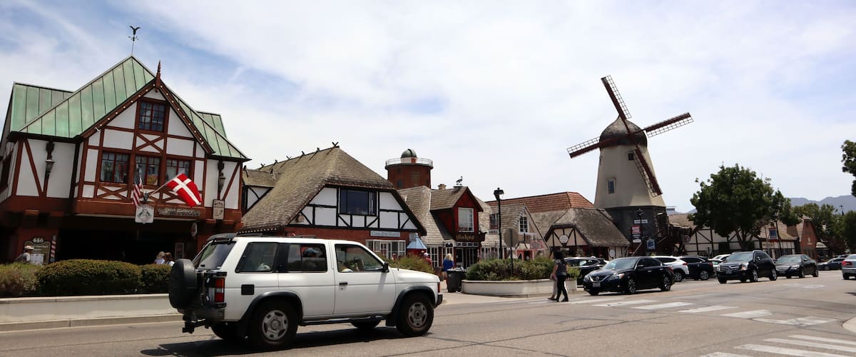 A white SUV driving on a street with traditional half-timbered buildings and a windmill.